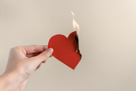 A paper red heart is burning in the hand of a woman on a beige background. Concept of unhappy love, separation, suffering.の写真素材