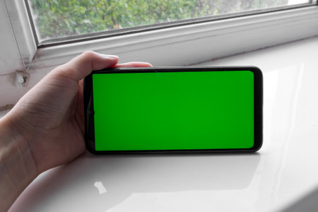 The woman's hand holds the smartphone in a horizontal position with a green screen on a window. Chrome key. Mock up.の写真素材