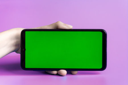 The woman's hand holds the smartphone in a horizontal position with a green screen on a pink background. Chrome key. Mock up.の写真素材