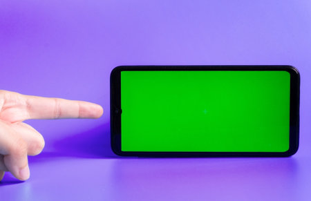 Smartphone in a horizontal position with a green screen on a purple background and a finger pointing at it. Chrome key. Mock up.の写真素材