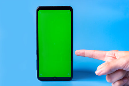 Smartphone in an upright position with a green screen on a blue background and a finger pointing at it. Chrome key. Mock up.の写真素材