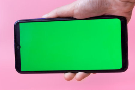 The woman's hand holds the smartphone in a horizontal position with a green screen on a pink background. Chrome key. Mock up.の写真素材