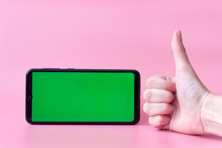 A smartphone in a horizontal position with a green screen on a pink background and a hand with a thumb up. Chrome key. Mock up.の写真素材