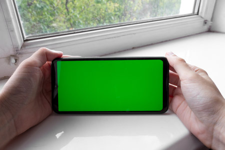 The woman's hands holds the smartphone in a horizontal position with a green screen on a window. Chrome key. Mock up.の写真素材