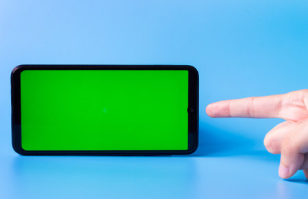 Smartphone in a horizontal position with a green screen on a blue background and a finger pointing at it. Chrome key. Mock up.の写真素材