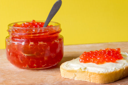 Red salmon caviar in a jar and on a butter sandwich. yellow background.の写真素材