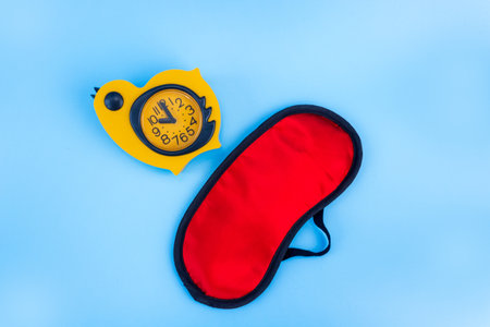 Red sleep mask with yellow alarm clock on a blue background. Insomnia, rest and healthy sleep concept. flat lay.の写真素材