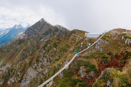 Two tourists go on a suspension bridge high in the mountains against a background of clouds.の写真素材