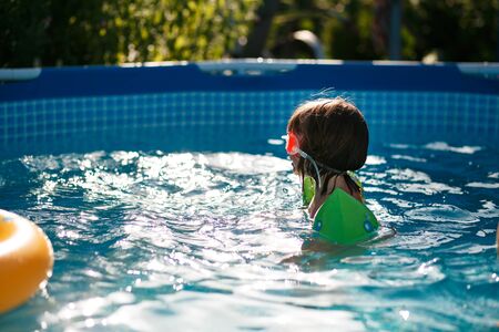 A child of three years old swims in a frame pool in the hot summer on the street in a mask and arm...