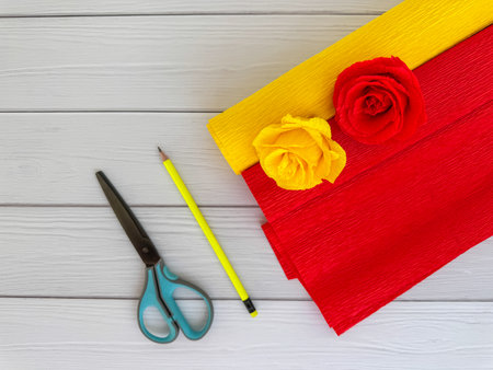 Red and yellow crepe paper rolls with handmade flowers, scissors, and pencil arranged on light wooden background. Perfect for DIY projects, crafts, and creative decorations.の写真素材