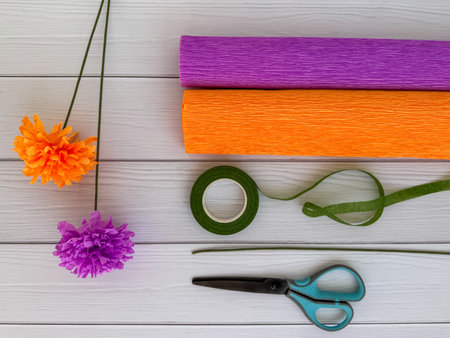 Handmade crepe paper flowers in purple and orange with matching rolls, green floral tape, scissors, and wire on light wooden background. Perfect for DIY crafts, creative projects, party decorations, and handmade gifts.の写真素材
