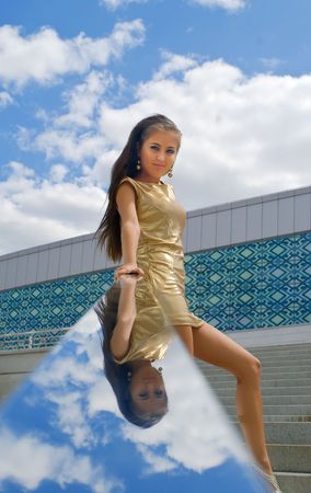 The girl in a golden dress at building steps against blue cloudy sky with reflexion in a ladder handrailの写真素材