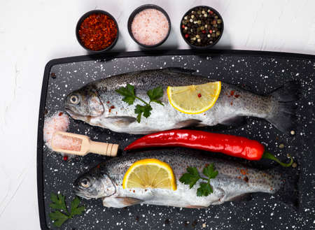 Fresh rainbow trouts on board on dark stone board with salt, pepper, lemon and rosemay ingredients....