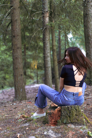 Back view of a young woman sitting in the forest, wearing jeans and a fashionable open-back top with...