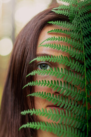 Close-up of a young woman's face seen through the slits of a fern leaf, creating a natural frame and...