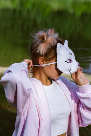 Teen girl in cat mask with pink eyes adjusts mask outdoors. Pink hoodie, crop top, messy bun with pink highlights. Quadrobics subculture, playful youth identity, expressive lifestyle.の写真素材