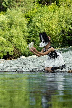 An enigmatic girl, part of the quadrobics subculture, interacts with nature. She kneels by a tranquil river, wearing a unique skull mask, gently cupping water in her hands in a lush forest setting.の写真素材