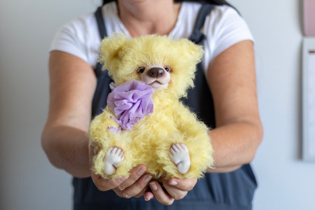 A woman's hands gently hold a fluffy yellow teddy bear, adorned with a delicate purple fabric embellishment. The meticulously sewn collectible bear radiates warmth and skilled artistry.の写真素材