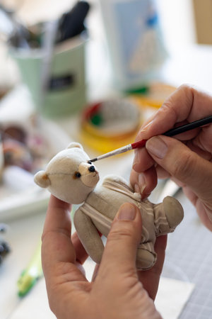 Close-up of an artist's hands meticulously painting the nose and mouth details on a small, handmade teddy bear. The bear is dressed in a tiny striped outfit, emphasizing the intricate craftsmanship.の写真素材