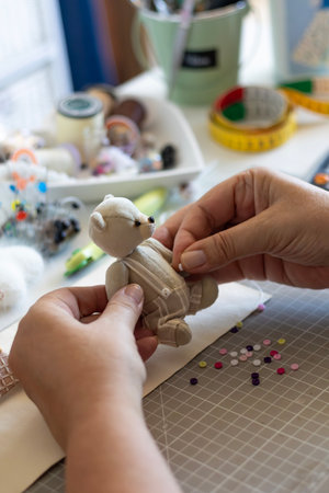 A close-up of hands carefully attaching a detail to a small, handmade teddy bear. The crafting table, filled with sewing accessories, highlights the intricate process of creating unique soft toys.の写真素材