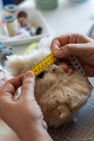 Crafter's hands precisely measure a cute, furry plush toy with a yellow tape measure, making a handmade stuffed animal.の写真素材