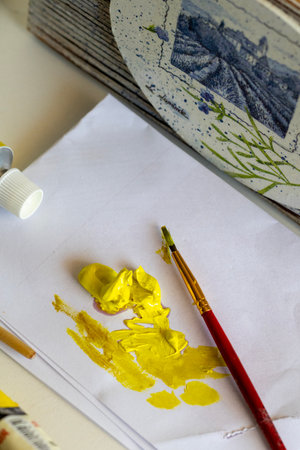 Vibrant yellow paint is squeezed onto white paper, accompanied by a paintbrush, signaling the start of a creative art project. A decorative plaque with a landscape painting adds an artistic backdrop.の写真素材
