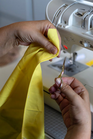 Close-up: Hands with scissors trim thread from yellow fabric being sewn. Sewing machine visible.