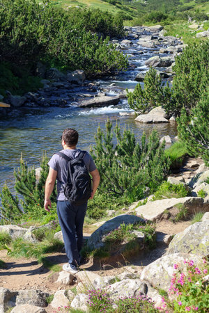 A man with a backpack hikes along a rocky path beside a clear mountain river in Pirin National Park. The lush green landscape and flowing water offer a serene natural escape.の写真素材