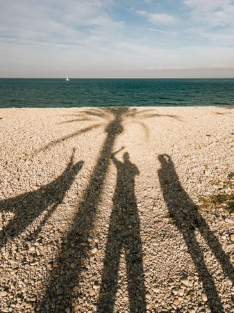 Shadows of three friends standing on pebble beach with arms raised, sailboat sailing on sea in background, sunny day with blue sky, white clouds. Three friends standing on rocky shoreline. Shadowsの写真素材