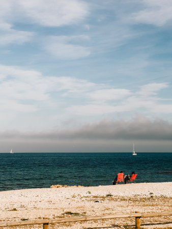 Couple relaxing on red chairs at rocky shoreline, overlooking calm blue waters with sailboats drifting against cloudy background, embodying peaceful seaside momentの写真素材