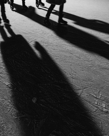 Long shadows stretching across an ice rink, capturing the lively scene of people skating on a sunny winter day, highlighting the striking black and white contrastの写真素材