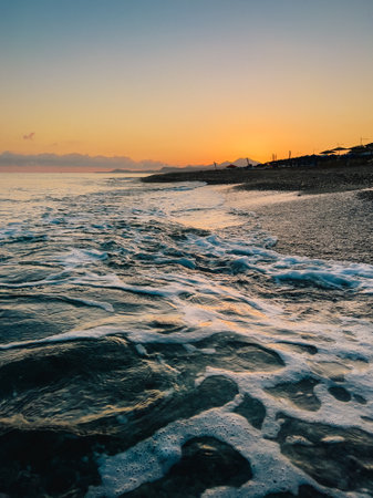 Soft waves lapping rocky shoreline during vibrant sunset along Cretan coastline, capturing serene Mediterranean summer evening atmosphereの写真素材