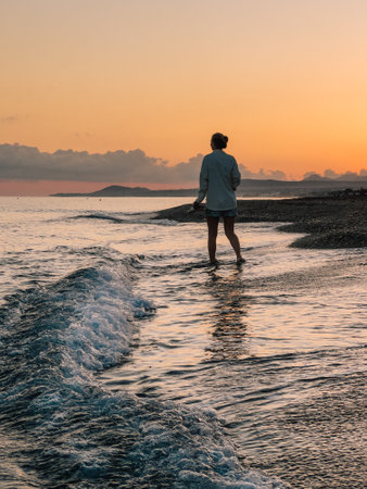 Barefoot woman walking coastal shoreline during golden sunset, experiencing serene landscape with warm orange sky and delicate wispy clouds, embodying peaceful solitudeの写真素材