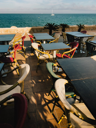 Seaside restaurant terrace featuring empty tables, vibrant chairs, sailboat gliding across blue horizon with calm ocean backdropの写真素材