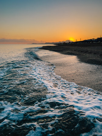 Gentle ocean waves washing over rocky shoreline, vibrant orange sunrise illuminating distant mountainous landscape with serene coastal sceneryの写真素材