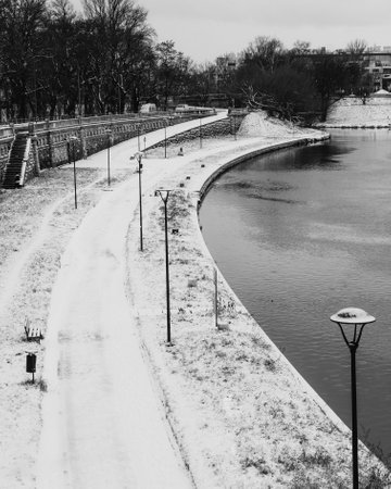 Snow covered promenade lining the river in winter, featuring street lamps, benches, and trash cans, captured in striking black and white image that evokes tranquility and solitude. promenade in winterの写真素材