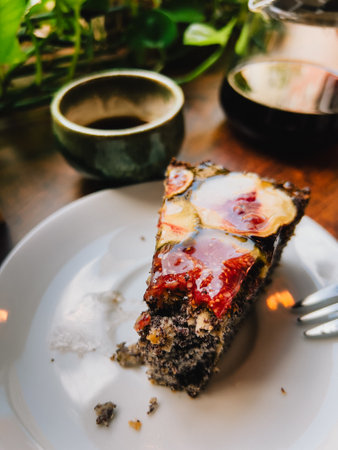 Poppy seed cake with fig slices and berry or fruit glaze on a white plate, partially eaten with a fork, accompanied by a cup of coffee or tea and a coffee pot on a wooden table. First-person viewの写真素材