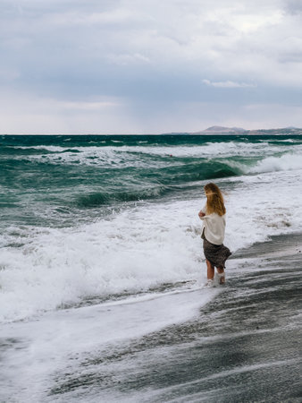 Young woman walking barefoot on beach enjoying stormy sea waves during cloudy day. Blonde woman wearing white sweater and long skirt walking barefoot along beach, embracing power of stormy sea wavesの写真素材