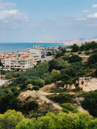 Lush greenery and a winding path dominate the foreground, leading the eye towards a picturesque cityscape nestled along the Greek coastline under a serene blue sky. overlooking coastal cityscapeの写真素材