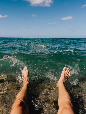POV shot of man relaxing on shore with his feet in transparent water of tropical sea, enjoying his summer vacation on sunny day. Travel and beach leisure concept. First-person view.の写真素材