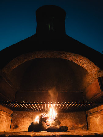 Small fire burning inside a barbecue grill at night, with the smoke coming out of the chimney and the dark blue night sky in the background. Wood burning inside a barbecue at night.の写真素材
