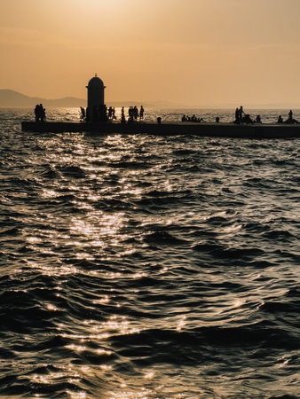 Tourists gathering along Zadar waterfront promenade at sunset, soaking in warm golden hour light as it dances on the waves of the Adriatic Sea, creating a picturesque scene. Tourists enjoying sunsetの写真素材