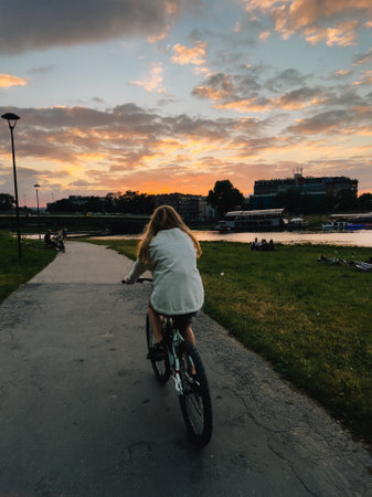 Young woman riding bicycle on paved path in park at sunset near river with boats and people relaxing on the grass, enjoying the colorful sky. Young woman riding bicycle in a park at sunset near riverの写真素材