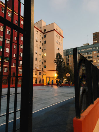 neighbourhood basketball court sunset lighting casting shadows as young players practicing viewed through weathered metal fencing between residential buildings. Urban basketball court, young peopleの写真素材