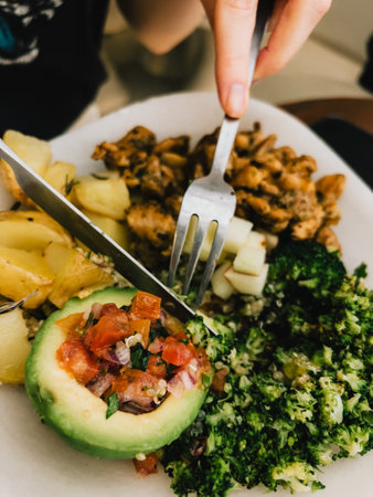 First-person view: Hands skillfully using cutlery while enjoying a healthy, balanced meal featuring broccoli, avocado, potatoes, and quinoa, highlighting fresh and nutritious ingredients.の写真素材