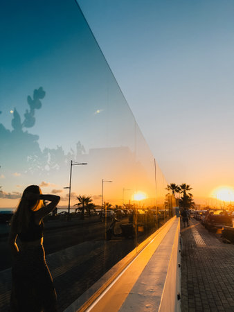 Golden sunset reflects on glass wall as woman gazes at glowing sky. Cityscape, palm trees, and mirror illusion enhance dreamy evening scene with elegant aesthetic and warm atmosphere. cinematic shotの写真素材
