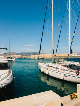 White sailboats and yachts gently rocking on tranquil turquoise waters of marina in Crete, Greece, under warm sunlight on clear summer day, create serene coastal scene. Sleek white sailboats rockingの写真素材