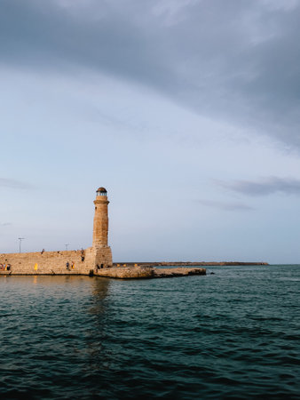 Venetian lighthouse overlooking historic Chania harbor, Crete, Greece, silhouetted against cloudy sunset sky with tourists nearby and surrounding Mediterranean waters. Venetian lighthouse, sea, Greeceの写真素材