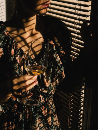Young woman wearing floral dress holding glass of white wine looking through venetian blinds casting shadows on her face and dress during sunset. Woman in floral dress sipping white wine, soft sunsetの写真素材