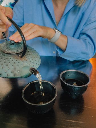 POV shot woman wearing blue shirt is pouring herbal tea from traditional japanese tetsubin teapot into two black textured cups on dark wooden table, creating cozy atmosphere. Herbal teaの写真素材